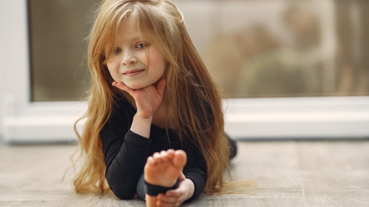 A young girl with long hair smiles while practicing yoga indoors, showcasing flexibility and joy.